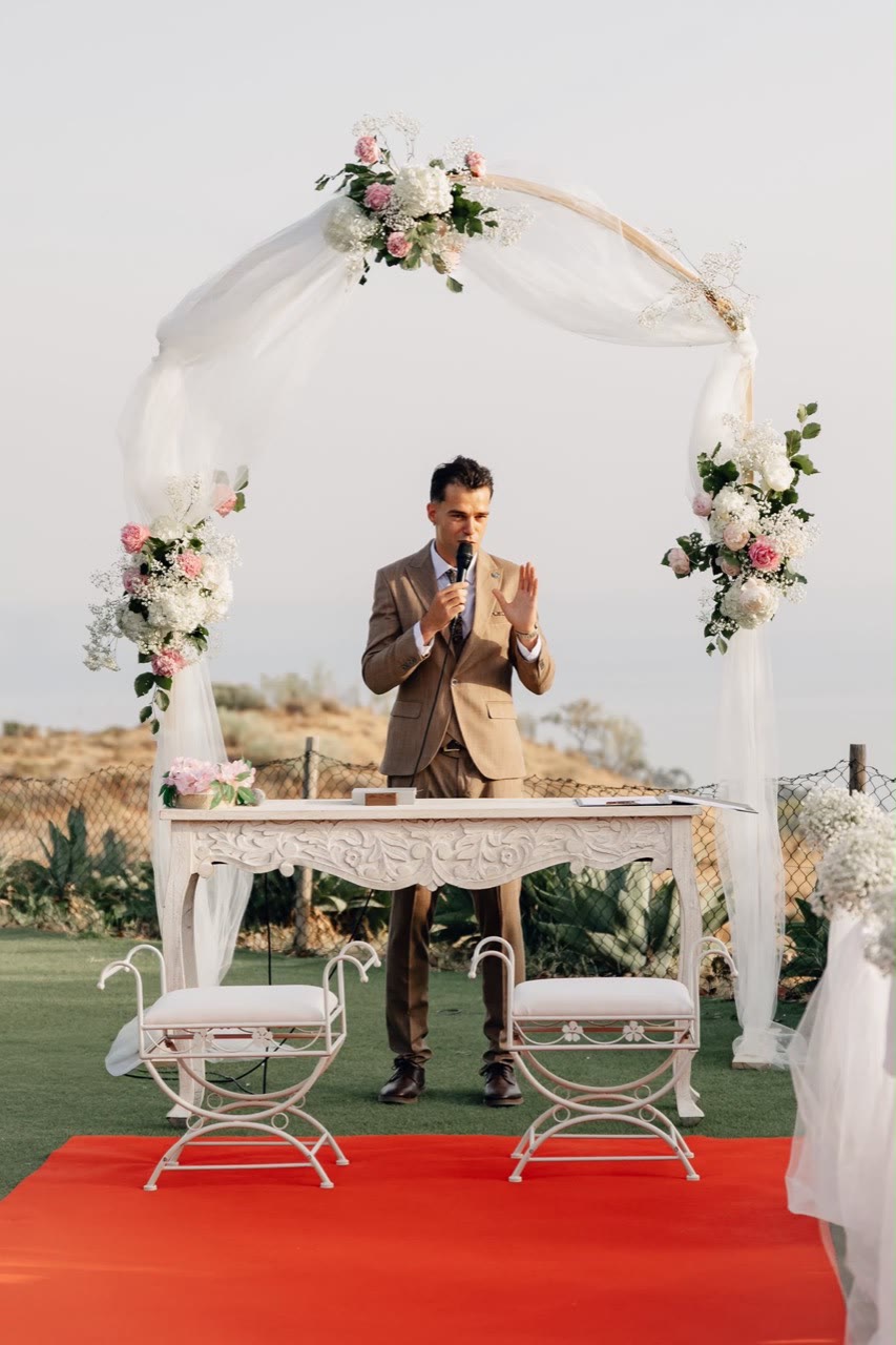 Vova standing beneath a floral ceremony arch in a tan suit
