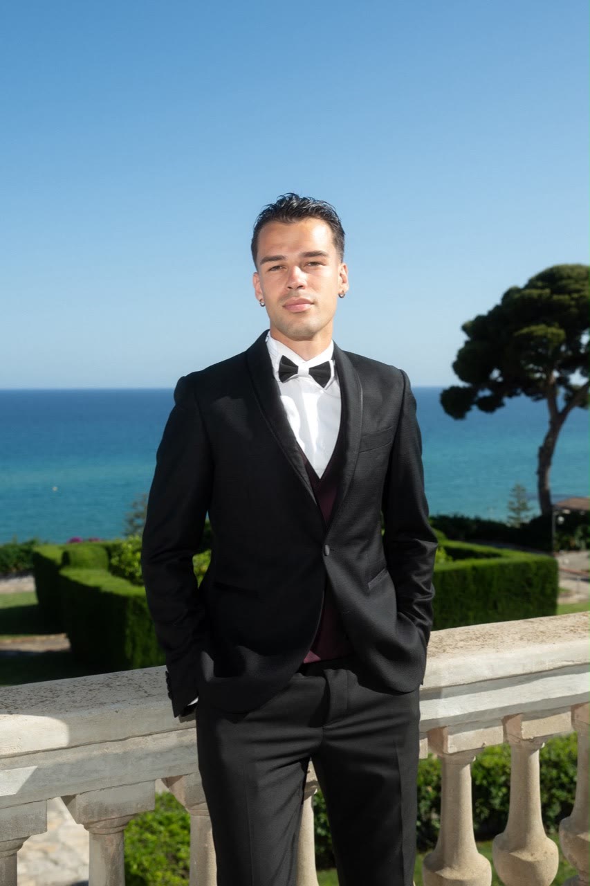 Vova in a black tuxedo standing in a garden above the sea